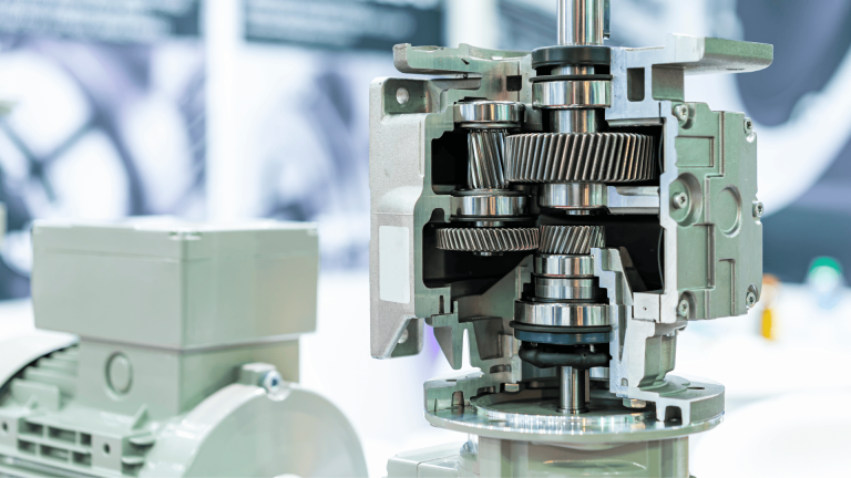 A transmissão mecânica industrial com a caixa de câmbio cortada está em exposição, revelando engrenagens helicoidais e rolamentos de metal. Um motor elétrico cinza está no plano de fundo desfocado.