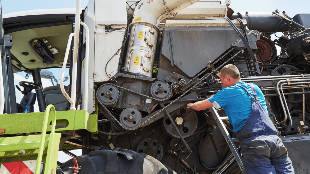 Mecânico realiza manutenção corretiva em colheitadeira agrícola, ajustando correias e correntes de transmissão com a máquina parada.