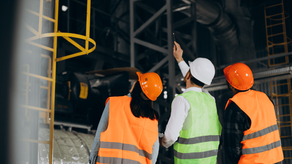 Equipe de engenheiros com capacetes e coletes de segurança analisando estruturas industriais, discutindo estratégias para aumentar a produtividade na manutenção.