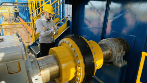 Profissional industrial utilizando EPI observa um acoplamento elástico para torque alto robusto amarelo e preto exposto entre eixos de maquinário, ilustrando a importância de analisar especificações técnicas ao entender como escolher o acoplamento ideal para garantir a eficiência e a segurança na transmissão de torque.