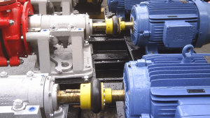 Dois conjuntos de equipamentos rotativos industriais montados em uma base metálica preta, mostrando bombas centrífugas acopladas a motores elétricos azuis por meio de eixos e acoplamentos amarelos e pretos. As bombas são vermelhas e prateadas, e as superfícies estão molhadas.