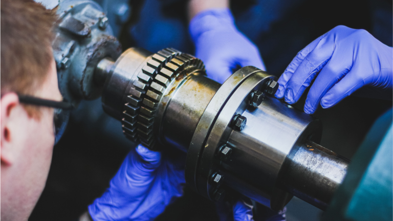 Close-up de um profissional com luvas azuis realizando a inspeção e ajuste manual de uma peça metálica industrial, etapa fundamental no processo de balanceamento de acoplamentos.