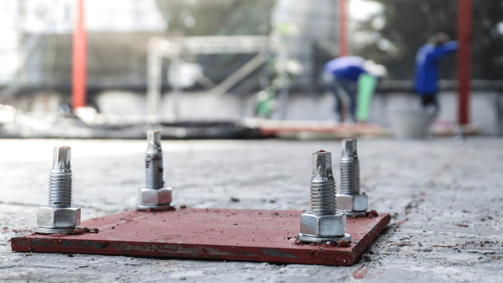 Close-up detalhado de quatro parafusos de ancoragem industrial com porcas e arruelas, fixando uma placa metálica vermelha sobre uma base de concreto em um canteiro de obras com operários ao fundo.