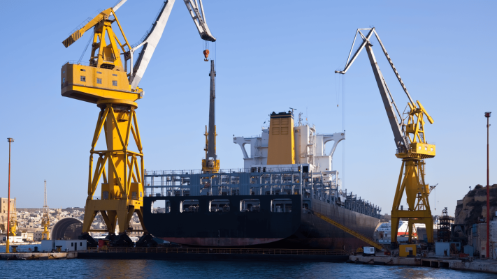 Um grande navio de carga atracado em um estaleiro sob um céu azul claro, ladeado por dois massivos guindastes amarelos, representando a infraestrutura de ancoragem industrial em operações portuárias de grande escala.
