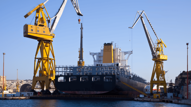 Um grande navio de carga atracado em um estaleiro sob um céu azul claro, ladeado por dois massivos guindastes amarelos, representando a infraestrutura de ancoragem industrial em operações portuárias de grande escala.