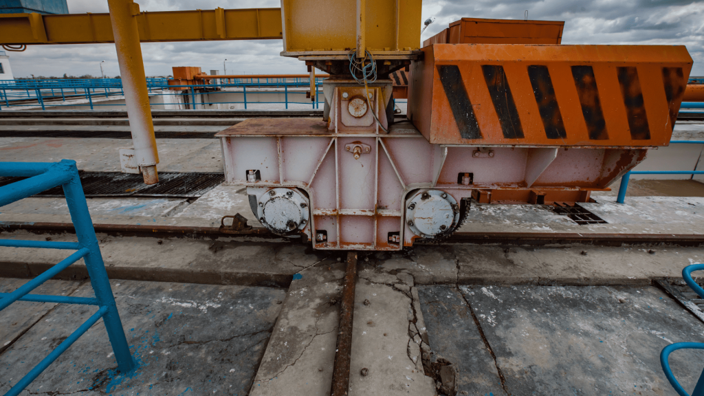 Close-up da base de um equipamento industrial pesado, mostrando as rodas metálicas e a estrutura de suporte sobre o trilho. O maquinário possui cores branca e laranja com faixas de sinalização, posicionado para garantir a fixação em trilhos em um pátio de concreto com marcas de desgaste.