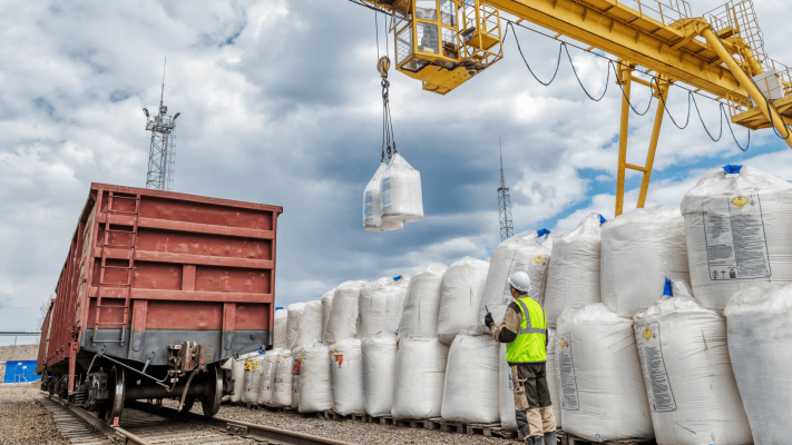 Operação logística externa com um guindaste de pórtico amarelo içando grandes sacos brancos (big bags) ao lado de um vagão de carga ferroviário. O guindaste utiliza um sistema de fixação em trilhos para movimentação segura, enquanto um trabalhador com colete de sinalização supervisiona a área.