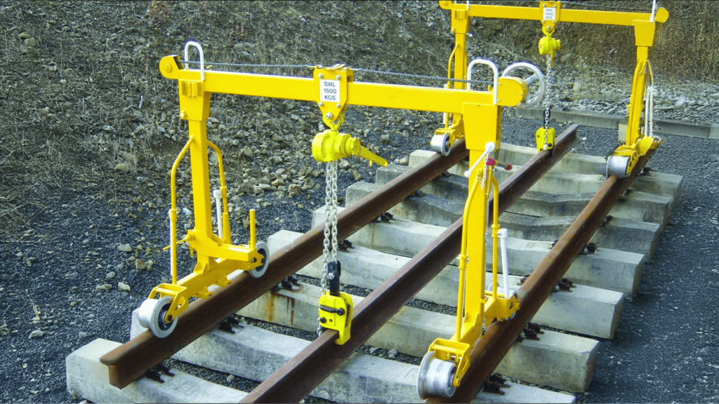 Duas pontes rolantes manuais amarelas posicionadas sobre trilhos ferroviários e dormentes de concreto. O equipamento é utilizado para o içamento e posicionamento preciso de trilhos, exemplificando um processo de fixação industrial em infraestrutura ferroviária.