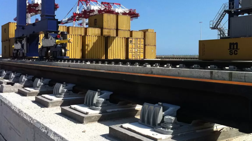 Detalhe em primeiro plano de trilhos de carga pesada fixados por grampos ajustáveis sobre bases de concreto em um porto. Ao fundo, veem-se contêineres e guindastes, destacando a robustez do sistema de fixação industrial e ancoragem industrial para operações logísticas de grande porte.