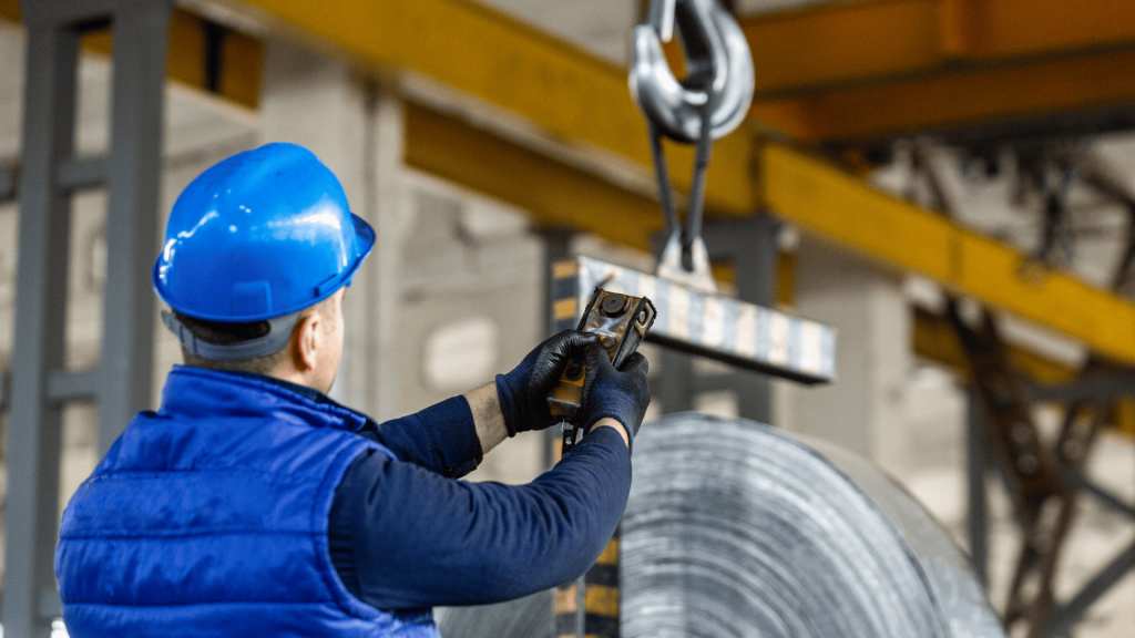 Operador industrial de costas, vestindo capacete azul e colete de proteção, manuseia um controle remoto para operação de carga em uma fábrica. A cena ilustra a segurança operacional em máquinas rotativas em um ambiente com bobinas de aço e estruturas de guindaste ao fundo.