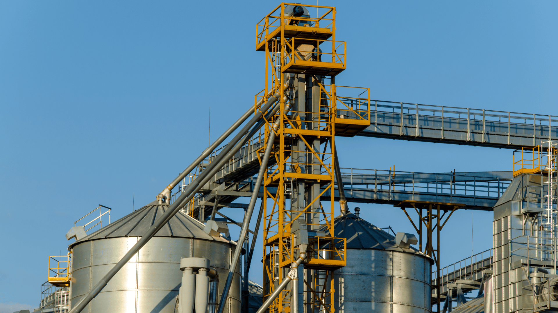 Vista lateral de uma unidade industrial de processamento com silos agrícolas metálicos e um elevador de canecas central em estrutura amarela. O sistema de acionamento no topo utiliza um acoplamento para elevador de canecas para transmitir potência ao transporte de grãos sob um céu azul.