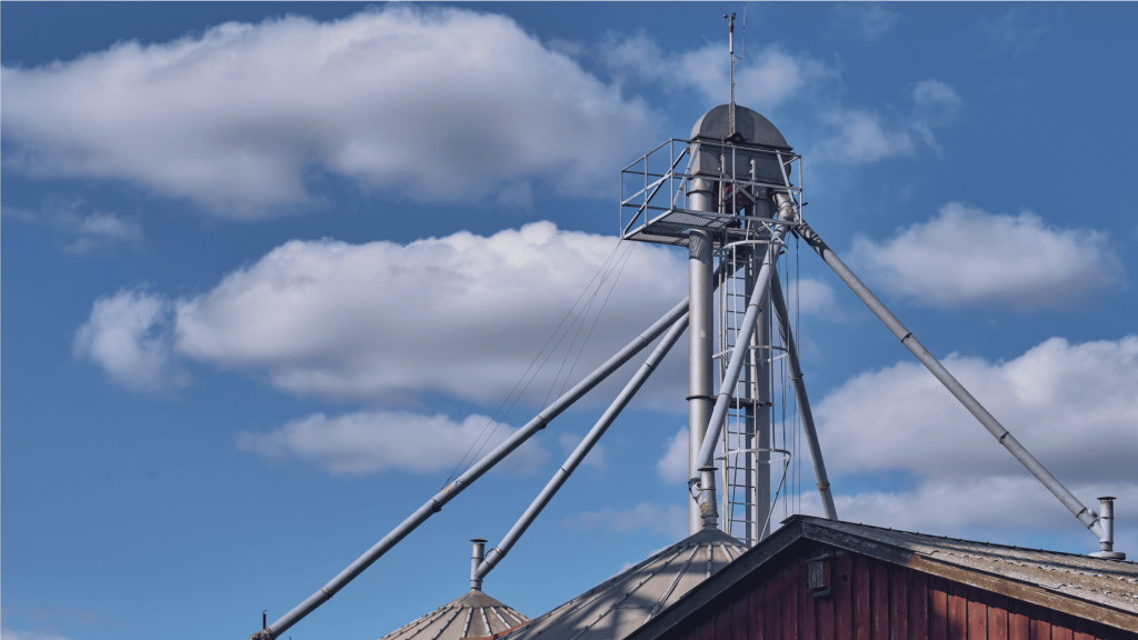 Elevador de canecas vertical metálico em uma fazenda, posicionado entre silos de armazenamento com nuvens ao fundo. A imagem destaca a parte superior do equipamento, onde o motor e o acoplamento para elevador de canecas garantem a movimentação contínua da carga.