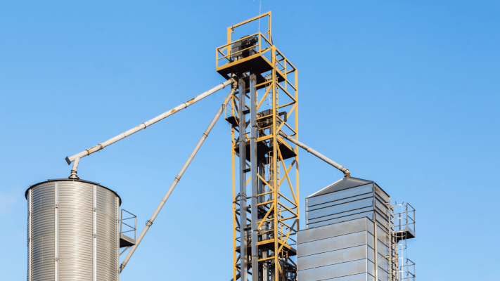 Estrutura de um elevador de canecas industrial amarelo conectando diferentes silos de metal. O conjunto motor no topo da torre é equipado com um acoplamento para elevador de canecas, peça fundamental para a eficiência e segurança operacional do sistema de elevação.