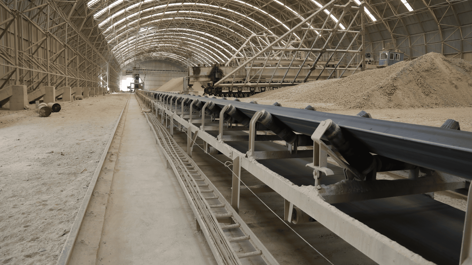 Uma visão interna de um enorme armazém de estrutura de aço em arco, com uma longa e vazia correia transportadora coberta de poeira estendendo-se ao longe. O piso de concreto e as pilhas de material de processamento estão cobertos de pó abrasivo. O ambiente fechado e empoeirado concentra as partículas, exigindo acoplamentos em ambientes extremos duráveis para resistir à contaminação e evitar paradas.