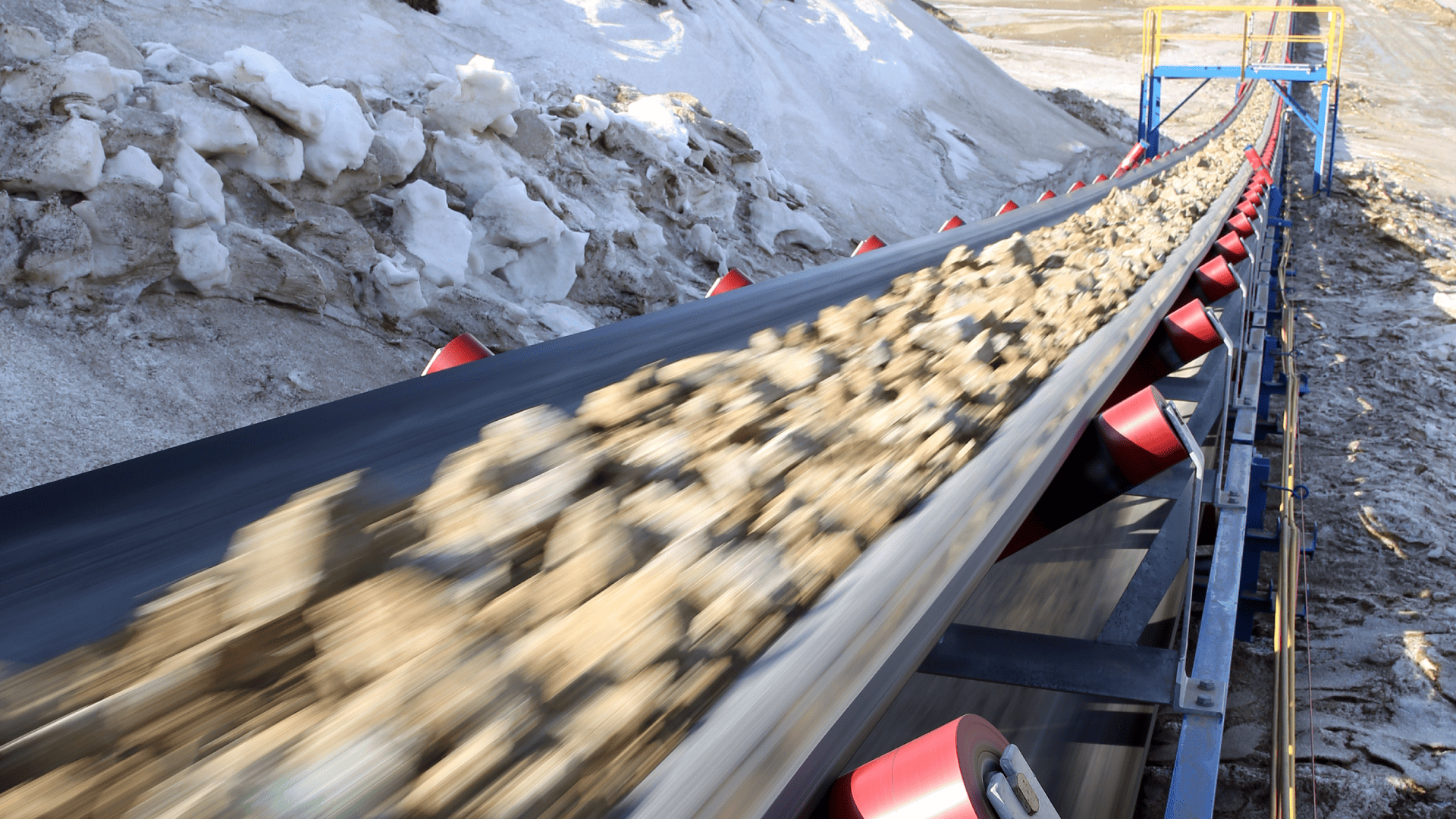Uma visão em perspectiva ao longo de uma correia transportadora de mineração que transporta rochas em alta velocidade. O equipamento está posicionado ao lado de grandes pilhas de neve e gelo sob um céu claro. Esta imagem destaca o desafio operacional de climas frios e a necessidade crítica de usar acoplamentos em ambientes extremos projetados para baixas temperaturas.