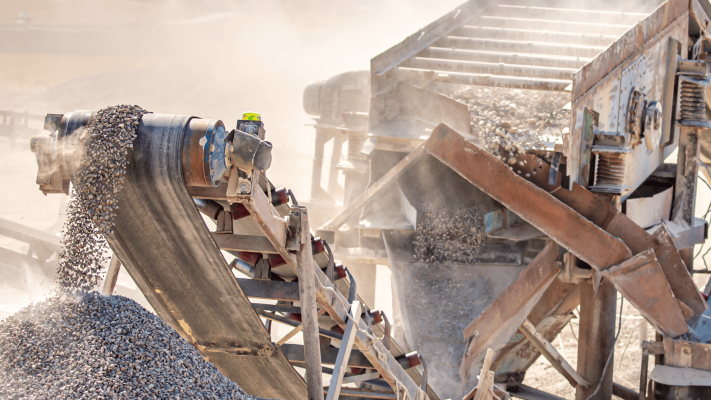 Um close de uma correia transportadora de pedreira coberta de poeira e ferrugem, despejando brita em uma pilha sob luz solar intensa. Uma nuvem densa de poeira envolve o motor e a estrutura, ilustrando o desgaste abrasivo e a vibração que os acoplamentos em ambientes extremos devem suportar para manter a operação em andamento.
