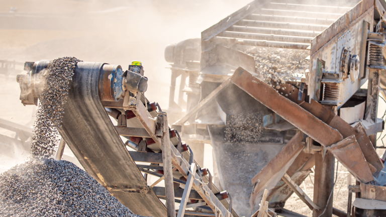 Um close de uma correia transportadora de pedreira coberta de poeira e ferrugem, despejando brita em uma pilha sob luz solar intensa. Uma nuvem densa de poeira envolve o motor e a estrutura, ilustrando o desgaste abrasivo e a vibração que os acoplamentos em ambientes extremos devem suportar para manter a operação em andamento.