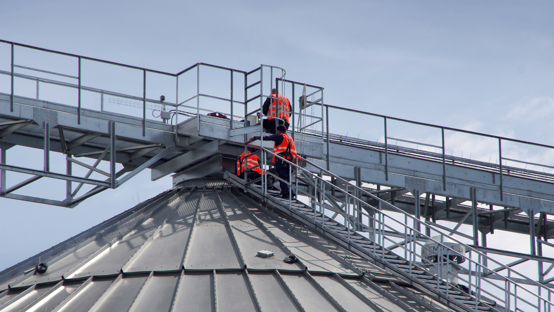 Três trabalhadores com coletes de segurança laranja realizam a manutenção na entressafra no topo de um silo metálico de grãos, sob um céu claro.