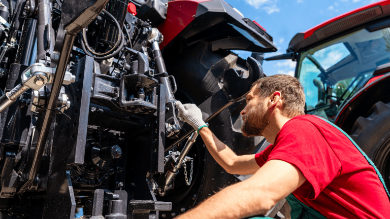 close-up de um mecânico inspecionando detalhadamente o engate e as engrenagens traseiras de um trator vermelho durante a manutenção na entressafra.