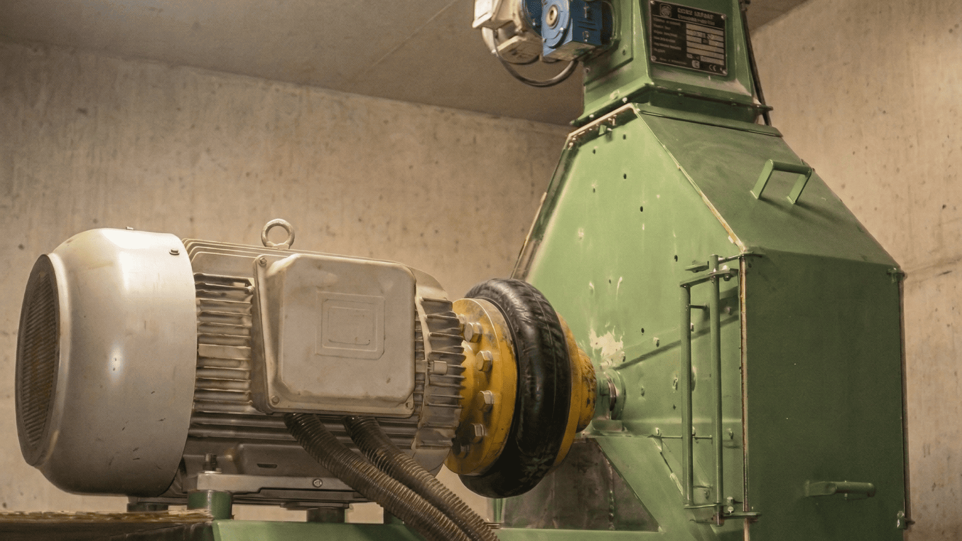 Close de um motor elétrico cinza e verde em um ambiente industrial, conectado a um moinho de martelo por meio de um acoplamento de disco flexível. Este é um exemplo de onde a seleção cuidadosa de acoplamentos para moinhos é crucial.