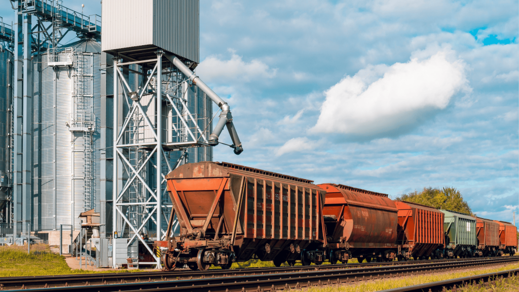 Vagões de trem posicionados para carregamento de grãos sob uma estrutura de silos metálicos, cujo sistema de movimentação de carga exige acoplamentos para equipamentos de transbordo de alta resistência.