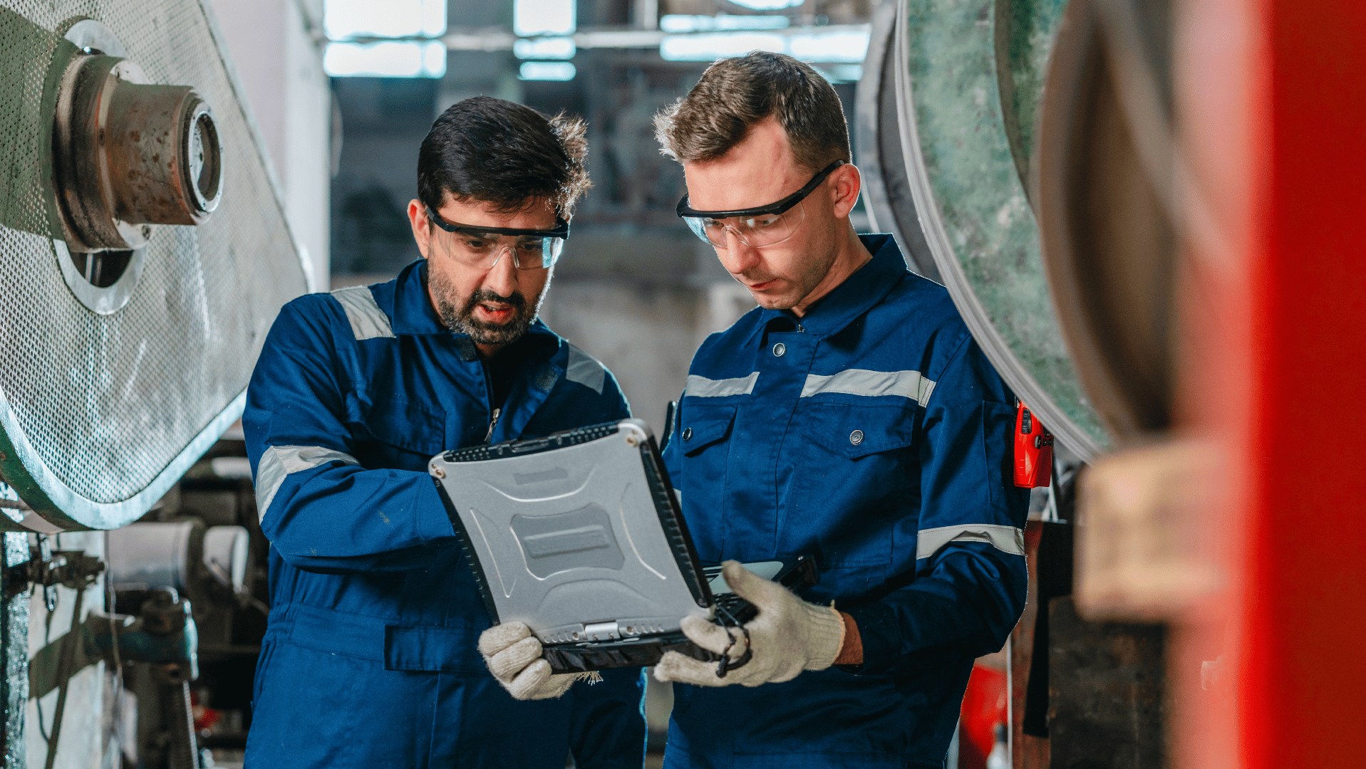 Dois técnicos de macacão azul e óculos de proteção analisam dados em um notebook robusto no chão de fábrica, monitorando o desempenho de máquinas conectadas via IIoT.