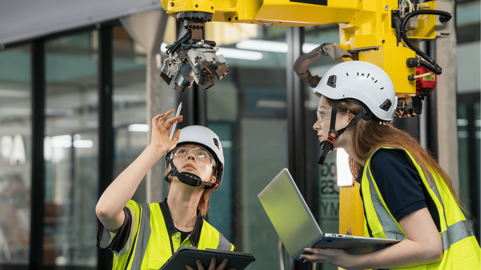 Duas engenheiras de capacete e colete refletivo inspecionam um braço robótico industrial usando tablet e notebook para coletar dados em um sistema IIoT.