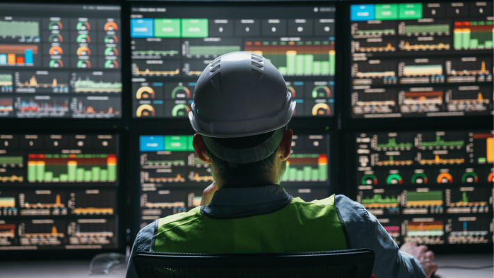 Visão traseira de um operador de capacete em frente a uma parede de telas com painéis digitais e gráficos complexos, supervisionando os indicadores de uma infraestrutura IIoT.