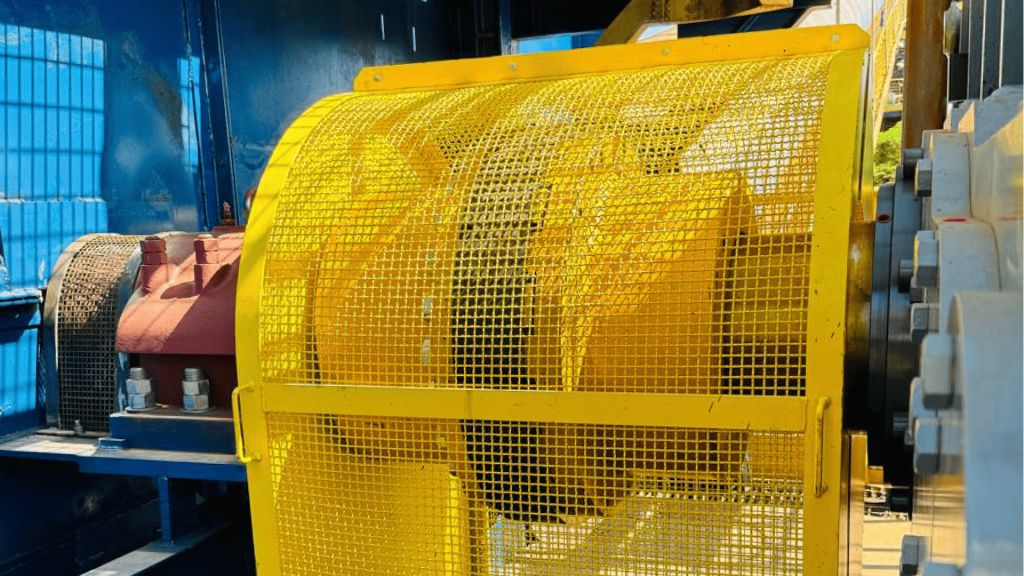 Vista inferior mostrando um acoplamento elástico para torque alto de grandes dimensões, protegido por uma grade de segurança amarela e montado entre eixos e rolamentos sob uma estrutura de plataforma industrial azul e amarela.