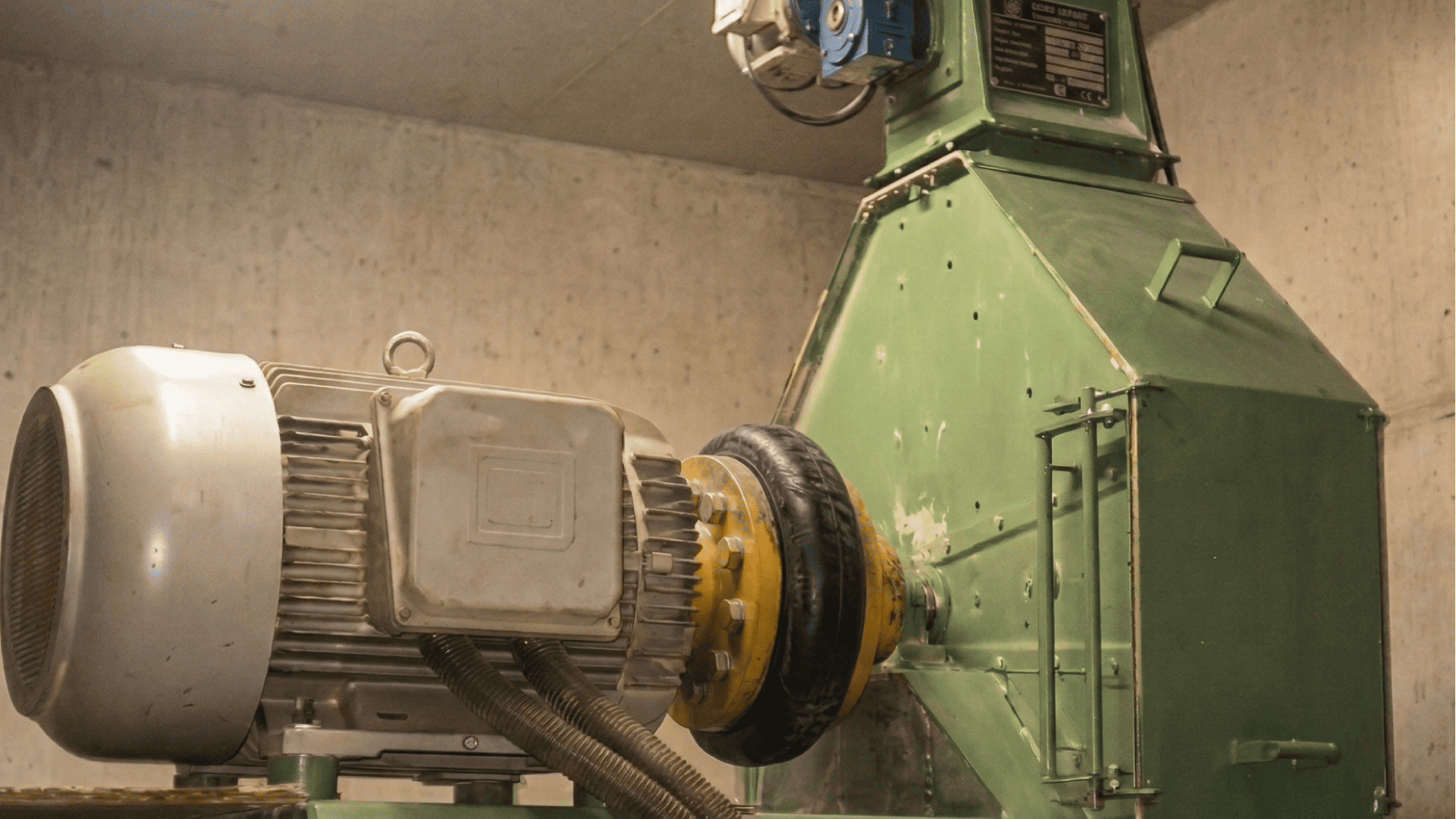 Instalação de acionamento industrial em uma sala de concreto, exibindo um motor elétrico acoplado a um grande invólucro de metal verde. O foco está no acoplamento flexível de pneu de borracha preta e amarela, que é um exemplo de acoplamentos para mineração para transmissão de potência e amortecimento de choque.