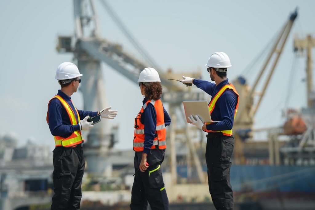 Três engenheiros com equipamentos de proteção individual, portátil e rádio a realizar a inspeção e análise de acionamentos críticos em operações portuárias no terreno, com guindastes e estruturas do terminal ao fundo.