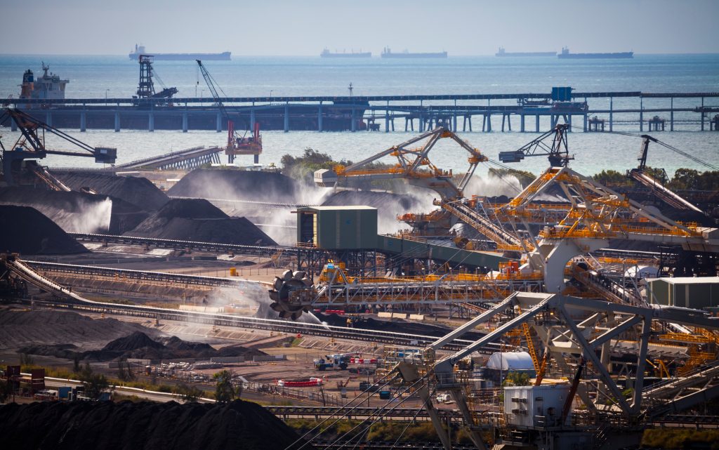 Vista aérea de um complexo terminal portuário de granéis, com maquinário para manuseio de materiais em operação, incluindo empilhadeiras-recuperadoras e transportadores de correia processando pilhas de carvão, com navios cargueiros ancorados ao fundo.